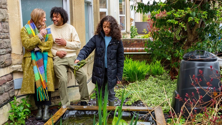 A family in their urban front garden by a pond. Two adults are leaning against the bay window, chatting with their hot drinks, while the young girl is playing by the pond